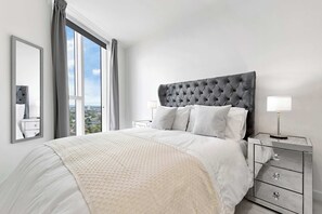 Ein modernes Schlafzimmer mit einem großen Bett, einem Nachttisch, einem Spiegel und Blick auf die Stadt durch das Fenster.