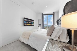 Ein modernes Schlafzimmer mit einem großen Bett, einem flachbildfernseher an der Wand, Blick auf die Stadt durch das Fenster und einem Nachttisch mit Lampe.