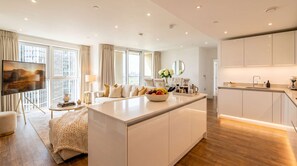 A modern living room with a large TV, a sofa, a coffee table, and a kitchen area with a counter and cabinets. - INSTAGRAM VIRAL SKY HIGH SANCTUARY With Gym and Exclusive Rooftop, Floor to Ceiling Windows (London)