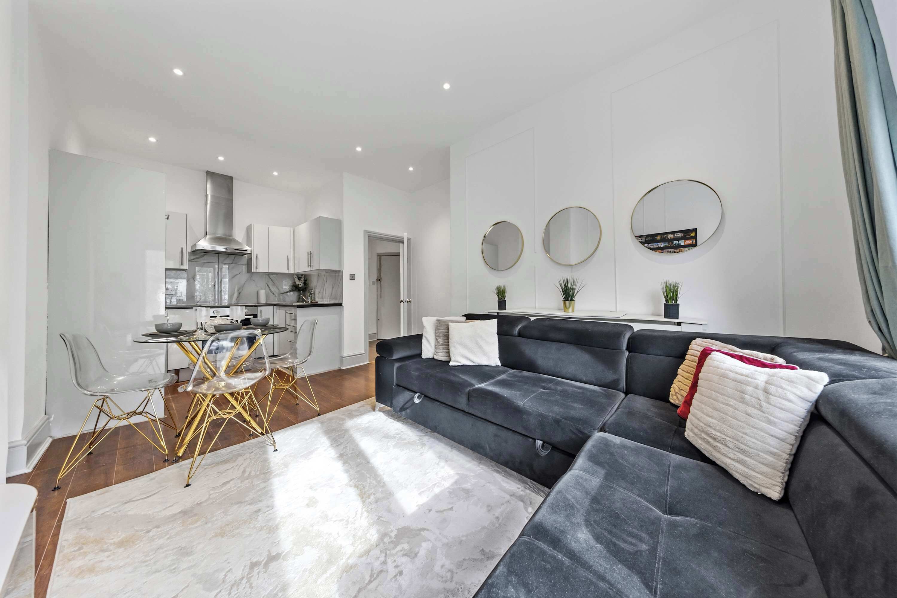 A modern living room with a black sofa, a dining area with a glass table and chairs, and a kitchen with white cabinets and stainless steel appliances.