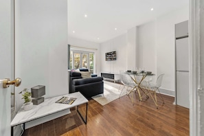 A modern living room with a sofa, a dining table with chairs, a kitchen area with a refrigerator, and a large window with a view of buildings. - Notting Hill Neighbour - 2BR Designer Stay (Greater London)