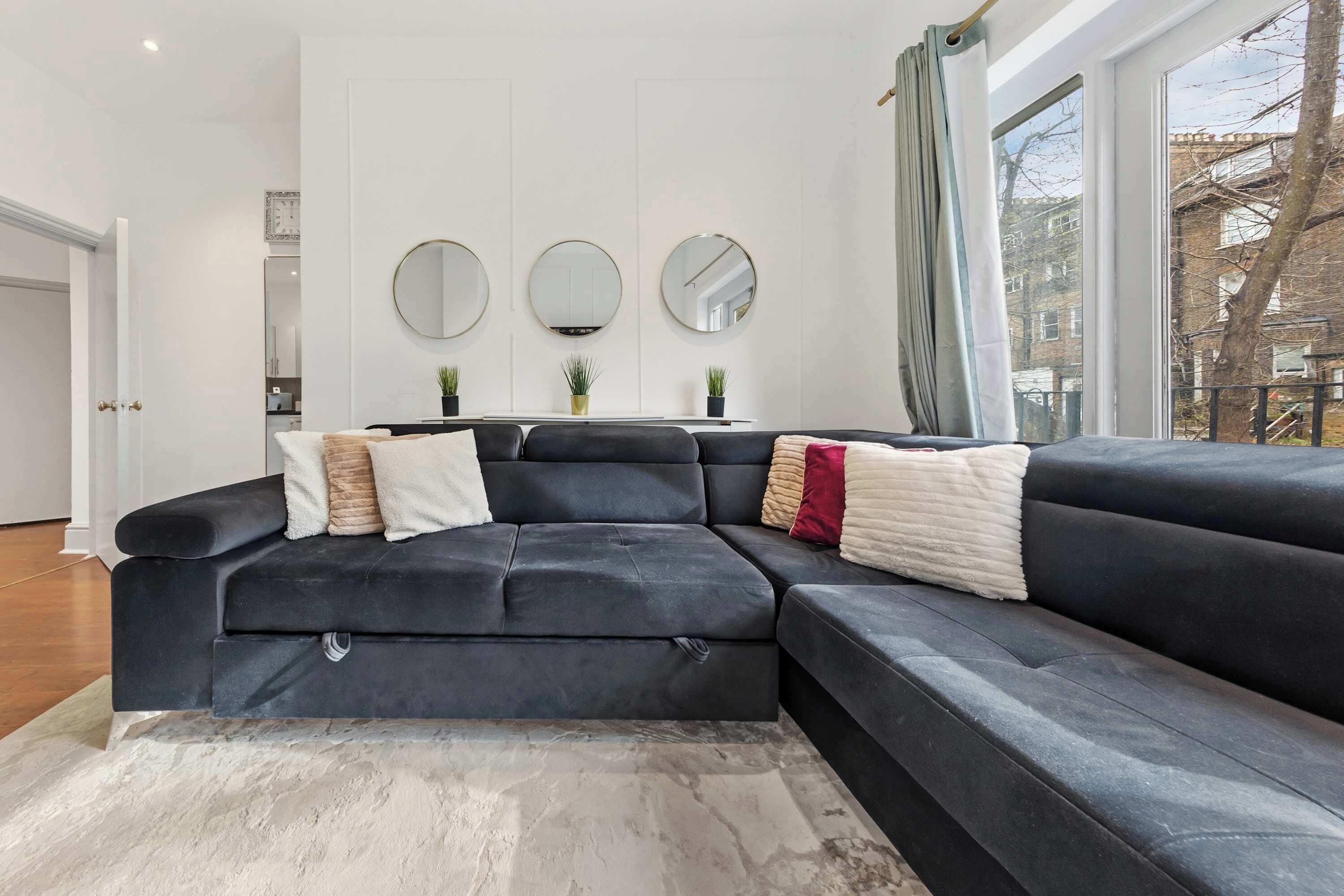 Ein modernes Wohnzimmer mit einem dunklen Sofa, drei runden Spiegeln an der Wand und einem Blick ins Freie durch ein Fenster.