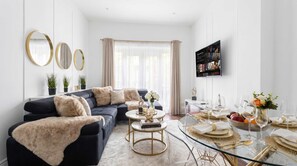 A modern living room with a sofa, coffee table, and a dining area set with glassware and plates. - Notting Hill Neighbour - 2BR Designer Stay (Greater London)