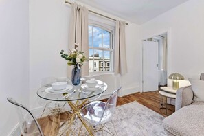 Ein modernes Wohnzimmer mit einem Glastisch, transparenten Stühlen, einer Vase mit Blumen und Blick auf Gebäude durch ein Fenster.