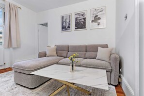 Ein Wohnzimmer mit einem Ecksofa, einem weißen Couchtisch mit goldenen Beinen und drei gerahmten Bildern an der Wand.