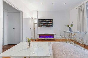 A modern living room with a flat-screen TV mounted on the wall, a coffee table, a floor lamp, and a dining area with a glass table and chairs. - Dream Open-View Balcony Apartment for 6 Guests, Moments from Harrods for Beautiful London Nights (Greater London)