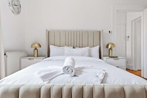 A neatly made bed with a rolled towel, two bedside tables with lamps, and a radiator. - Dream Open-View Balcony Apartment for 6 Guests, Moments from Harrods for Beautiful London Nights (Greater London)