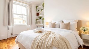 Una cama bien hecha con edredón blanco, una mesita de noche con lámpara y una ventana con cortinas.