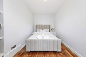 A narrow hotel room with a bed, bedside table, and a nightstand with a lamp. - Dream Open-View Balcony Apartment for 6 Guests, Moments from Harrods for Beautiful London Nights (Greater London)