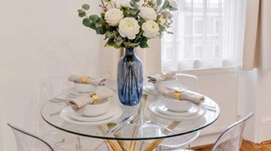 Una mesa de vidrio para comer con platos blancos, servilletas dobladas y un jarrón azul con flores blancas.