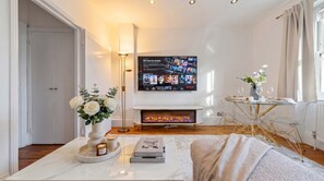 Una sala de estar moderna con chimenea, televisor montado, una mesa de centro con libros y velas, y un jarrón de flores.