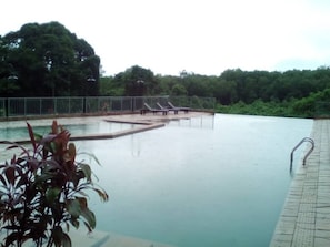 Una piscina con solárium alicatado, rodeada por una valla metálica y árboles al fondo.