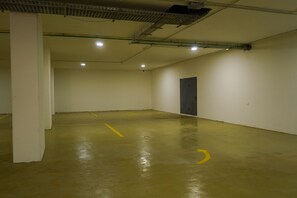Um amplo espaço de estacionamento coberto, com piso de concreto, vagas demarcadas e uma porta à direita.