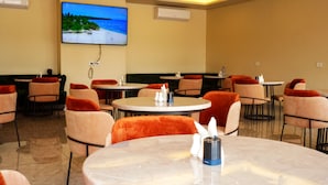 A hotel dining area with round tables, cushioned chairs, and a TV screen displaying a beach scene. - HOTEL PANORAMA (Kinshasa)