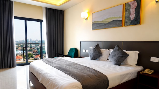 A hotel room with a large bed, a view of the city, a chair, and a nightstand with a phone. - HOTEL PANORAMA (Kinshasa)
