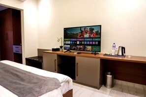 Habitación de hotel con cama, escritorio con computadora, televisor mostrando un partido de fútbol, cesto de basura y una botella de agua.