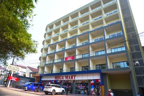 Un immeuble à plusieurs étages, avec un magasin « MEGA MART » au rez-de-chaussée, une enseigne « HOTEL PANORAMA » au-dessus, et un fourgon bleu garé devant.