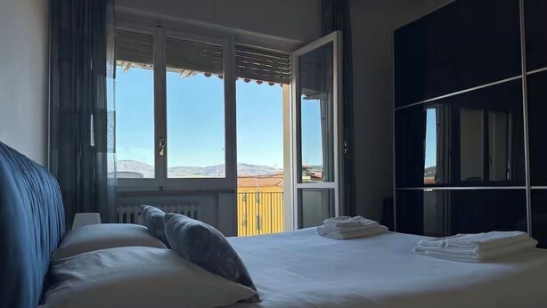 Een slaapkamer met een bed, een klein rond tafeltje, een groot raam met zonwering en uitzicht op buiten.