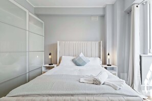 Ein ordentlich bezogenes Bett mit weißem Kopfteil, zwei Nachttischlampen und einem zusammengerollten Handtuch auf dem Bett.