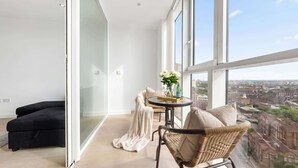 En balkong med en soffa, en flätstol, ett bord med en vas med blommor och utsikt över staden.