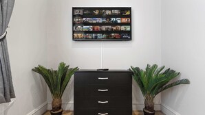 A black filing cabinet with three drawers, flanked by two potted plants, against a white wall with a mounted TV displaying various shows. - Cinematic Slice of HARRODS for 6 Hideaway Steps from Station (Greater London)
