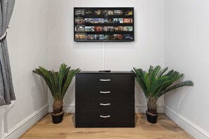 A black filing cabinet with three drawers, flanked by two potted plants, against a white wall with a mounted TV displaying various shows. - Cinematic Slice of HARRODS for 6 Hideaway Steps from Station (Greater London)