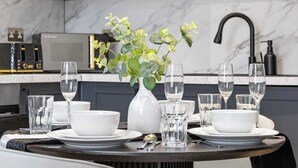 A dining table set with white plates, bowls, and glasses, accompanied by a vase with green foliage. - Cinematic Slice of HARRODS for 6 Hideaway Steps from Station (Greater London)