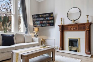 A living room with a sofa, a coffee table, a fireplace, a wall-mounted TV, and a window with curtains. - Cinematic Slice of HARRODS for 6 Hideaway Steps from Station (Greater London)