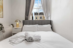 A neatly made bed with white linens and a rolled towel, a bedside lamp, and a view of buildings through a window with curtains. - Cinematic Slice of HARRODS for 6 Hideaway Steps from Station (Greater London)