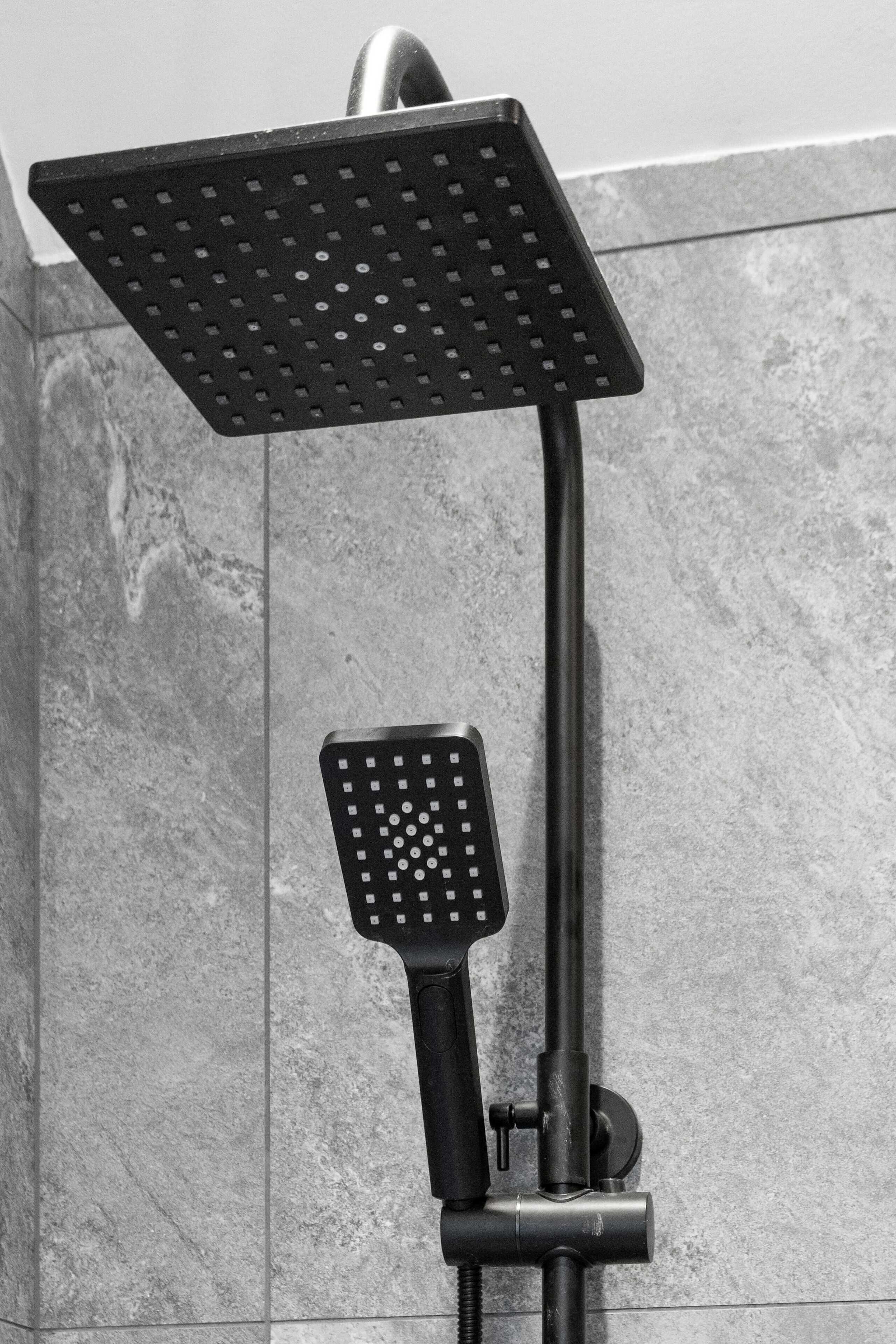 A showerhead with a square, perforated design, mounted on a vertical pipe, against a tiled wall.