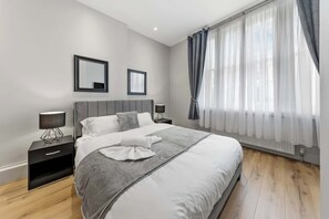 Un dormitorio moderno con una cama grande, dos mesitas de noche, una mesilla auxiliar, un espejo en la pared y cortinas translúcidas.