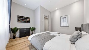 A modern bedroom with a large bed, a black dresser, two potted plants, a wall-mounted TV, and a framed picture. - Cinematic Slice of HARRODS for 6 Hideaway Steps from Station (Greater London)