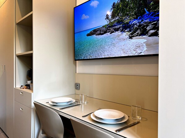 A modern dining area with a glass table, plates, cutlery, and a TV displaying a beach scene.