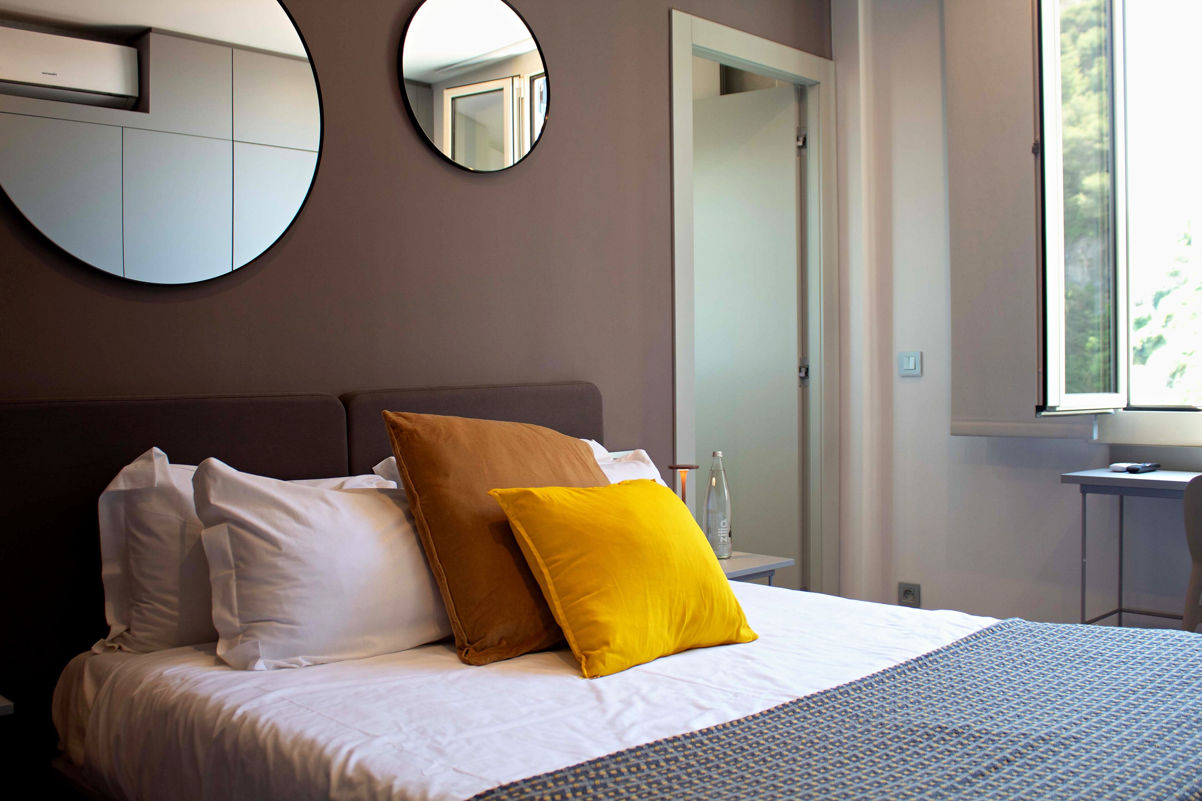A neatly made bed with white and brown pillows, a round mirror on the wall, and a window with a view of greenery.