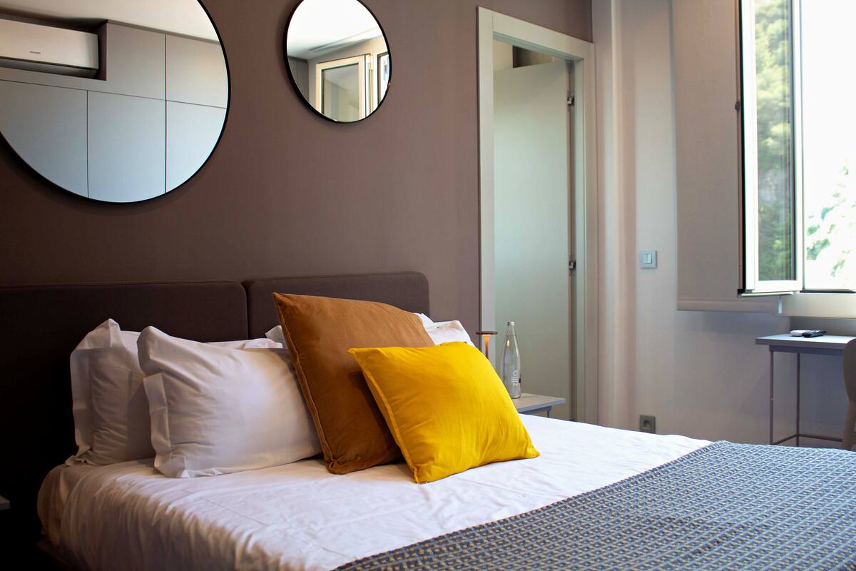 A neatly made bed with white and brown pillows, a round mirror on the wall, and a window with a view of greenery.