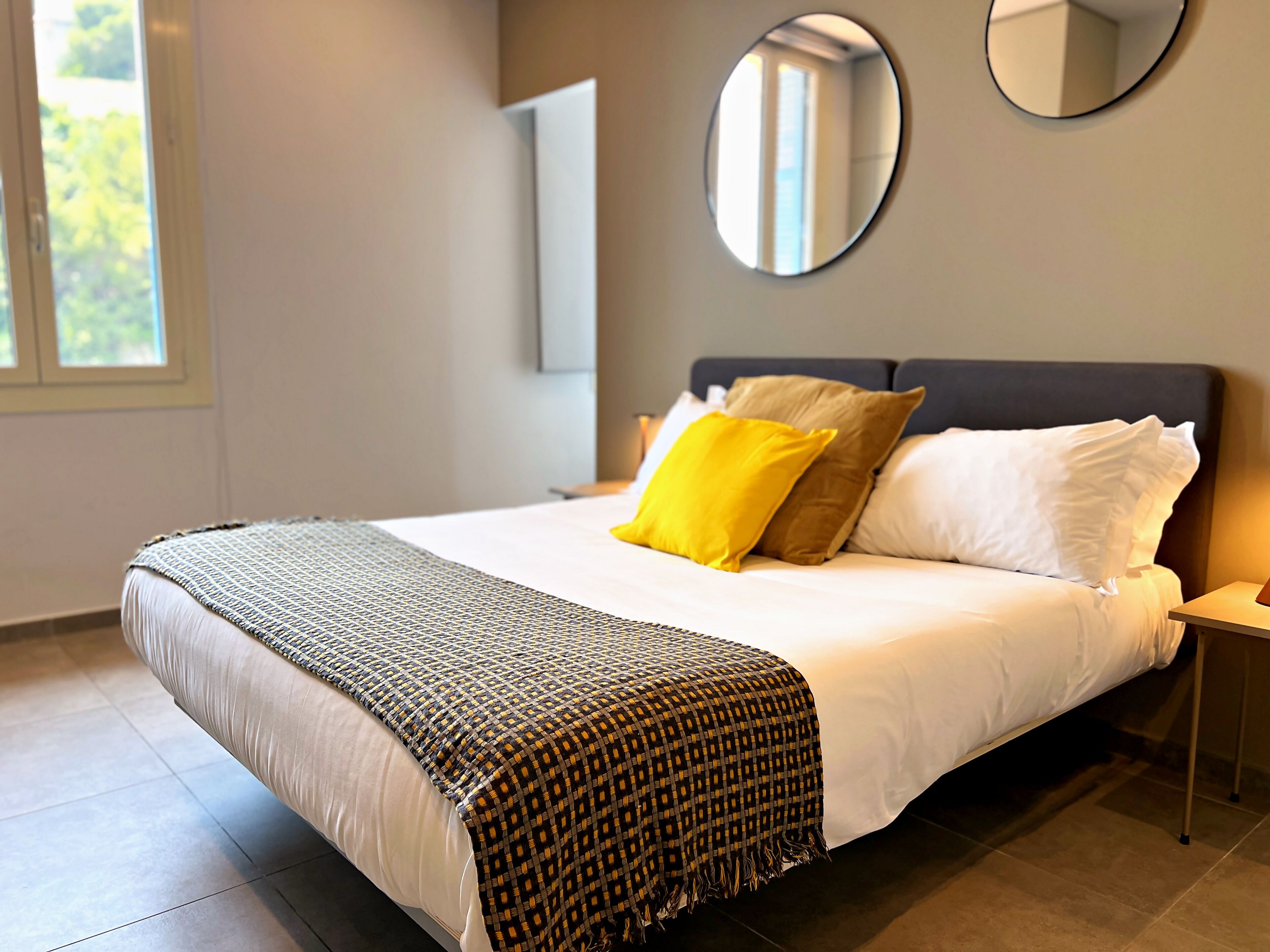 A neatly made bed with a patterned blanket, a bedside table with a lamp, and a round mirror on the wall.