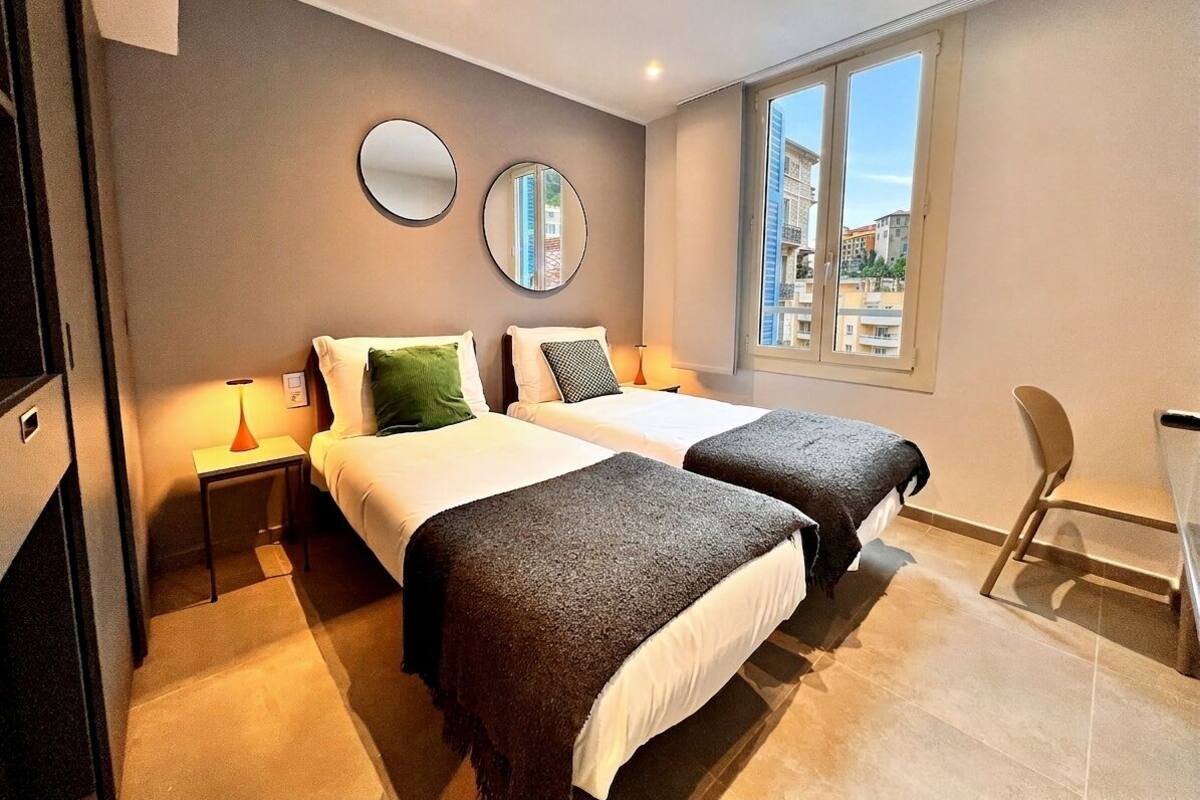 A hotel room with two beds, a window with a view, a small table with a lamp, and a chair.