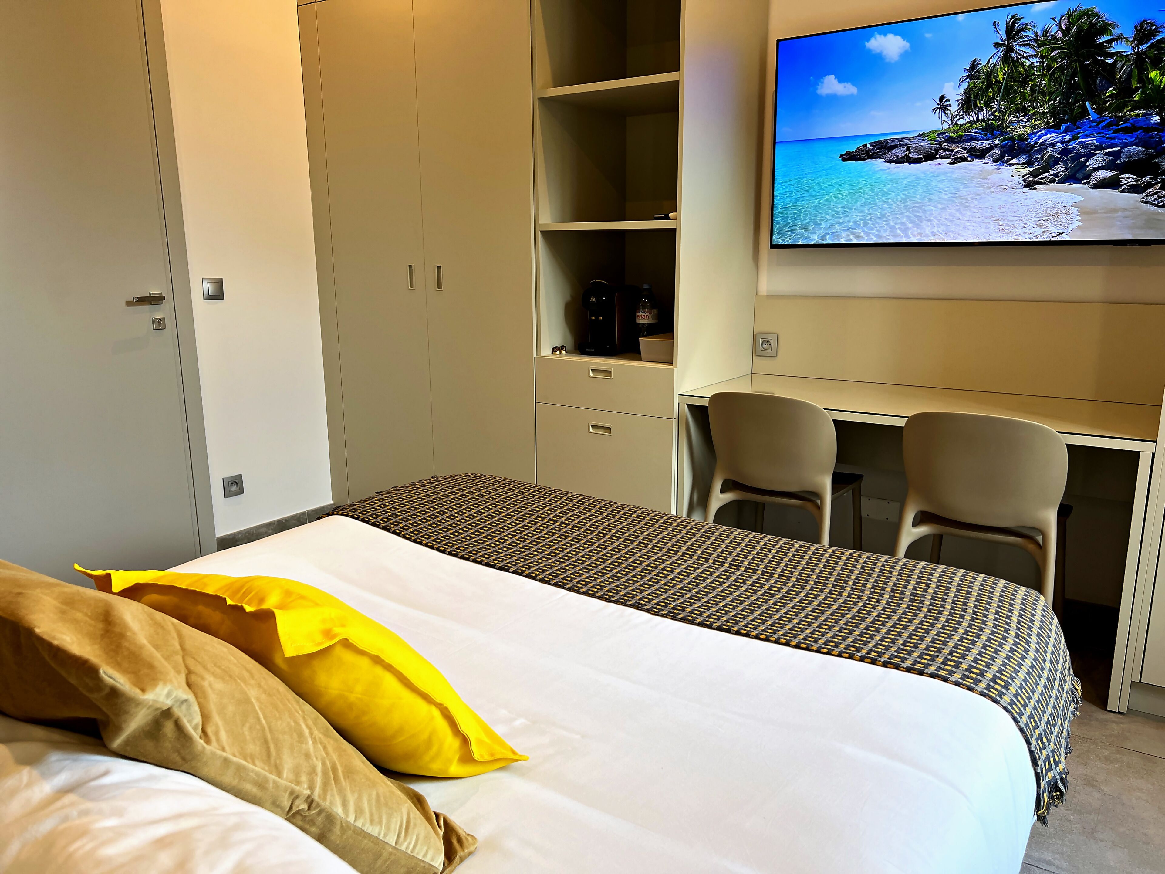 Ein Hotelzimmer mit einem Bett, einem Schreibtisch mit zwei Stühlen, einem Kleiderschrank und einem Fernseher, der eine Strandansicht zeigt.