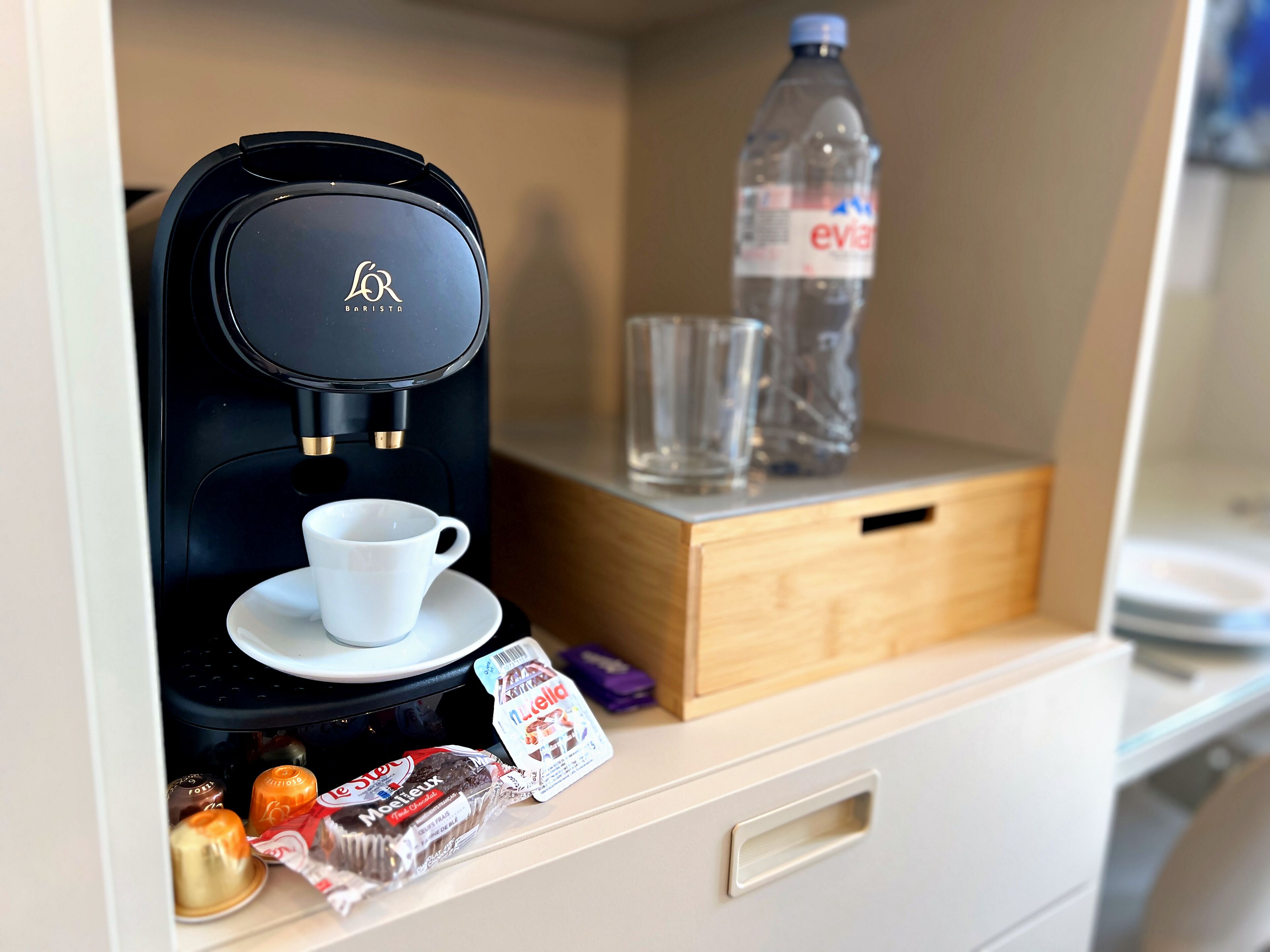 Une machine à café, un verre, une bouteille d’eau et quelques en-cas emballés sur un comptoir.