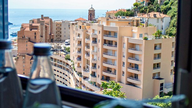 Vista de un paisaje urbano costero a través de una ventana, con edificios de varios pisos, balcones y un mar distante.