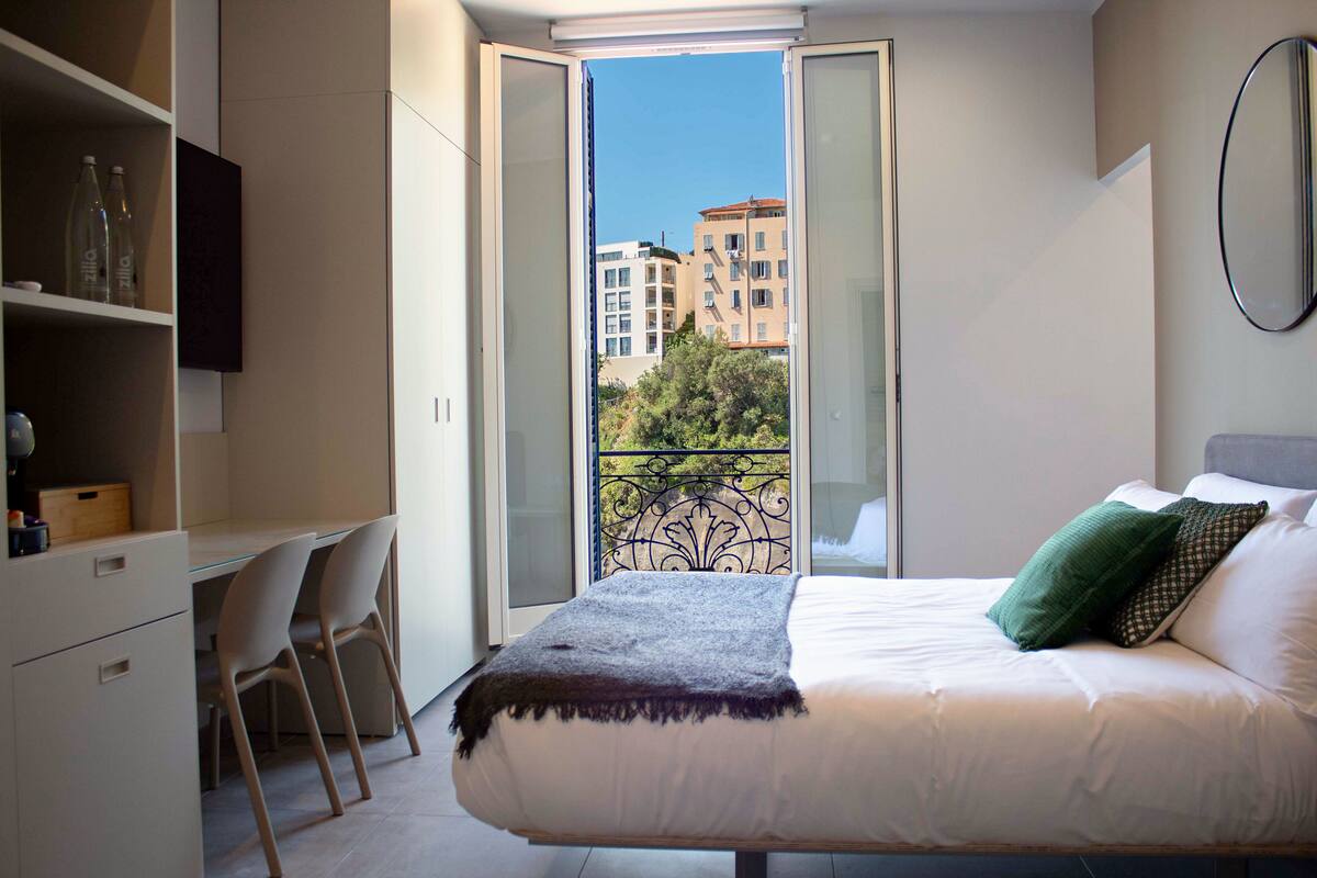 A bedroom with a bed, a desk, chairs, and a balcony with a view of buildings.