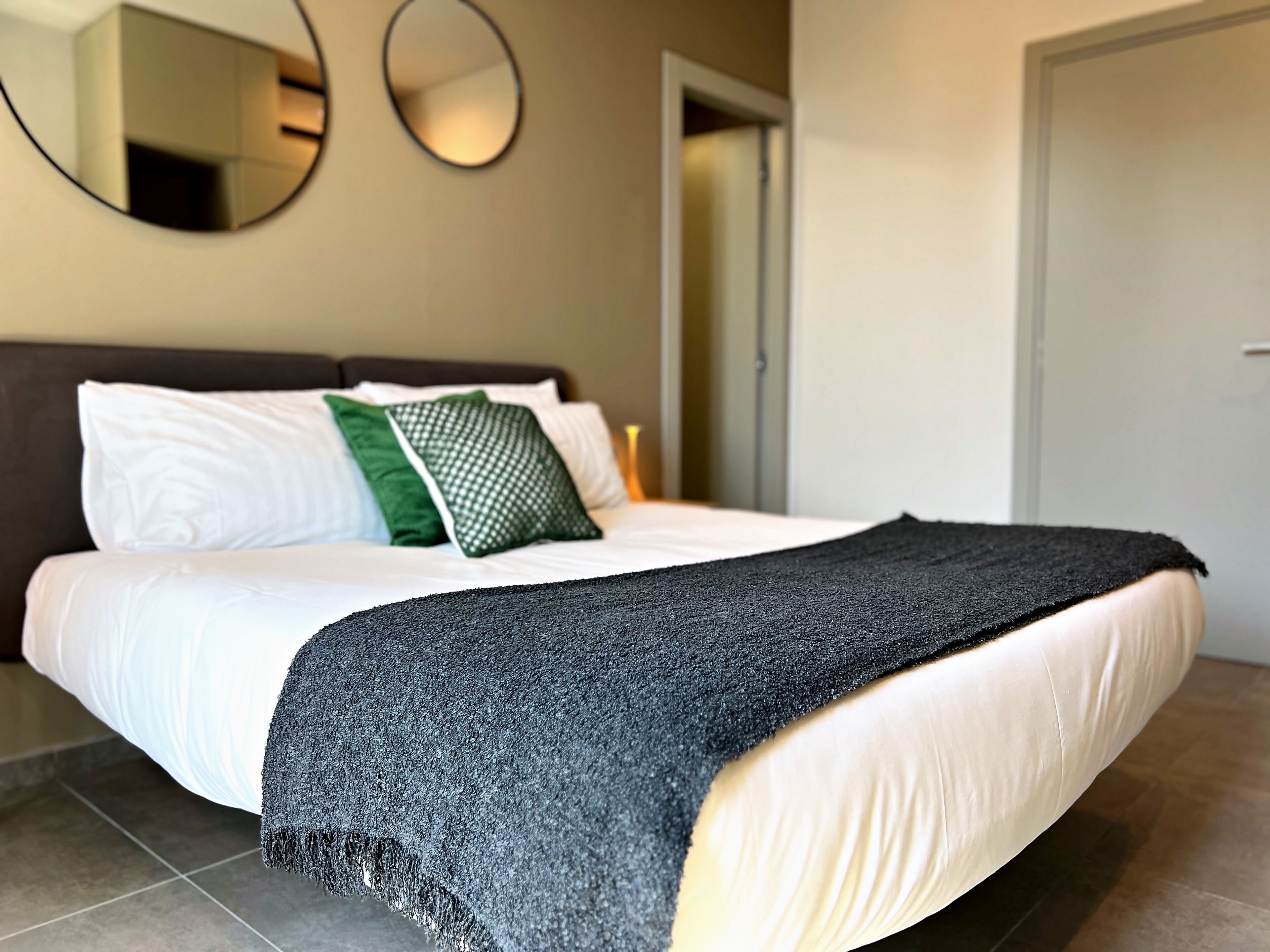 A neatly made bed with white linens, a dark grey blanket, and green and white pillows.