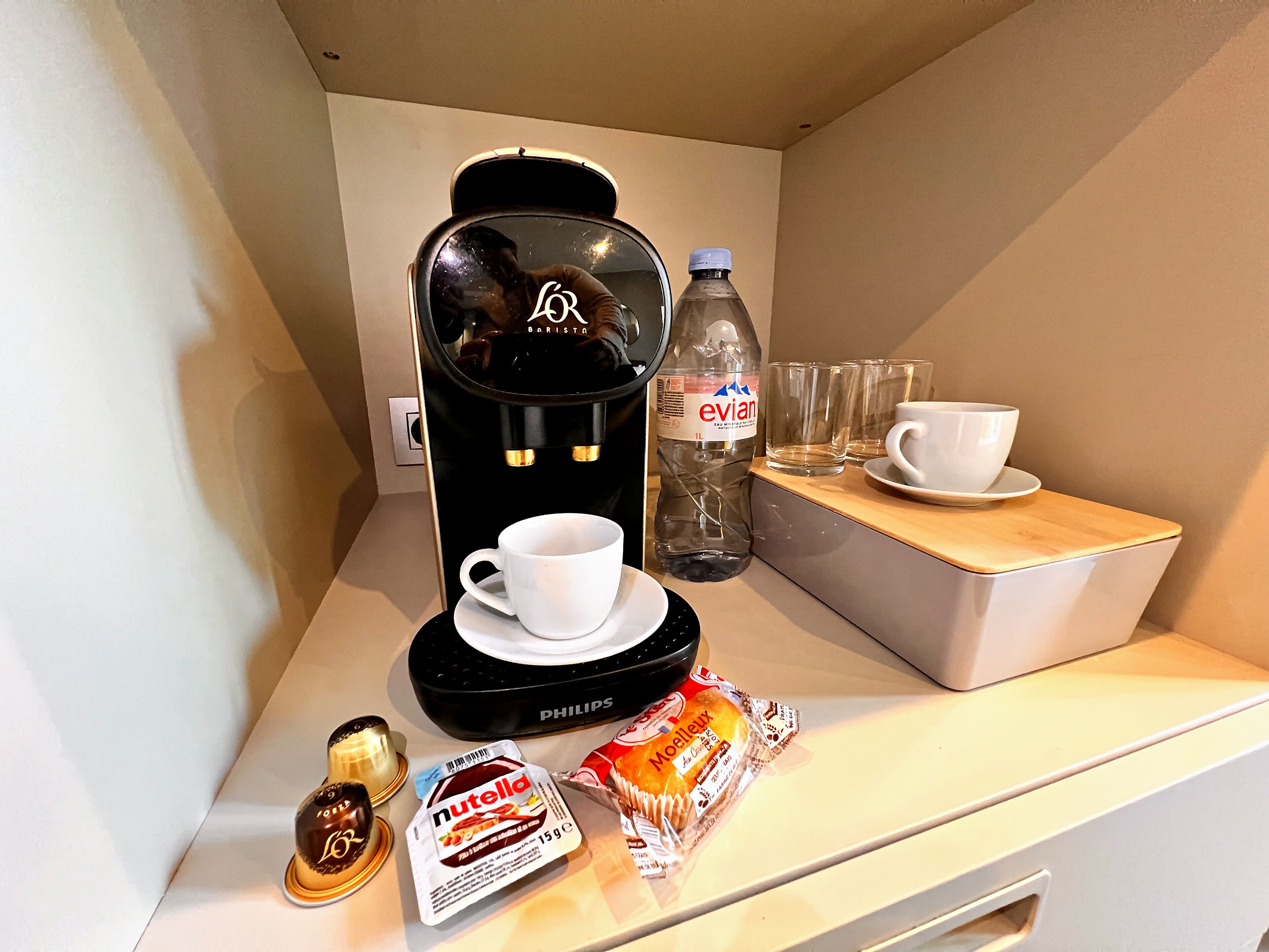 A coffee machine, a bottle of Evian water, two glasses, a cup, a Nutella bar, and a chocolate bar on a counter.