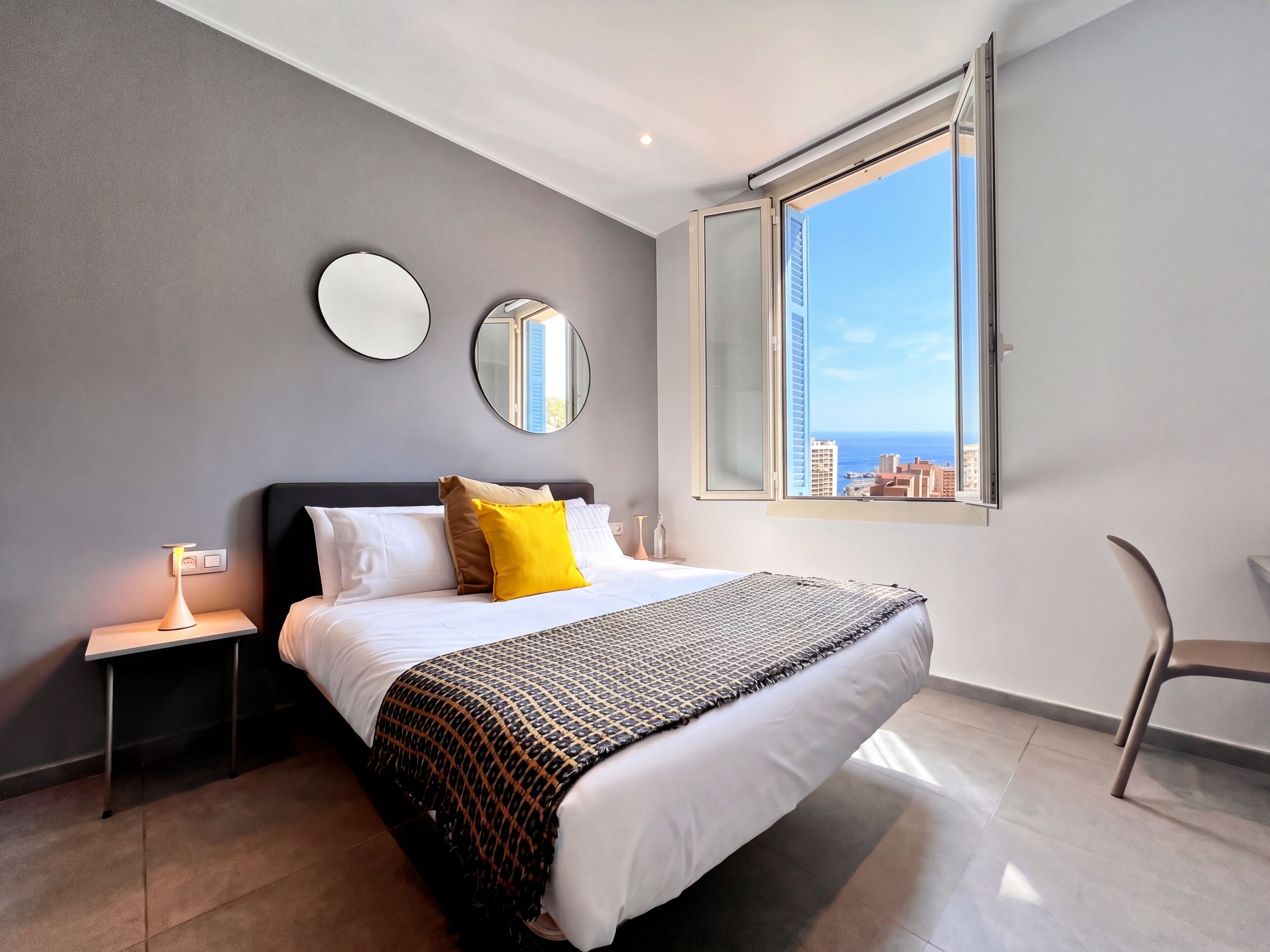 A modern bedroom with a bed, bedside tables, a chair, and a large window offering a city view.