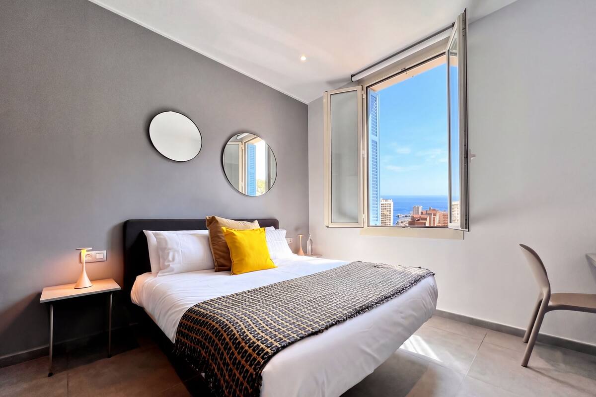 A modern bedroom with a bed, bedside tables, a chair, and a large window offering a city view.