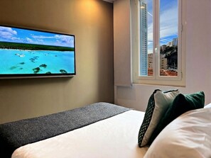 A hotel room with a bed, a television displaying a scenic view, a window with blinds, and a view of buildings. - My Residence d'Azur (Beausoleil)