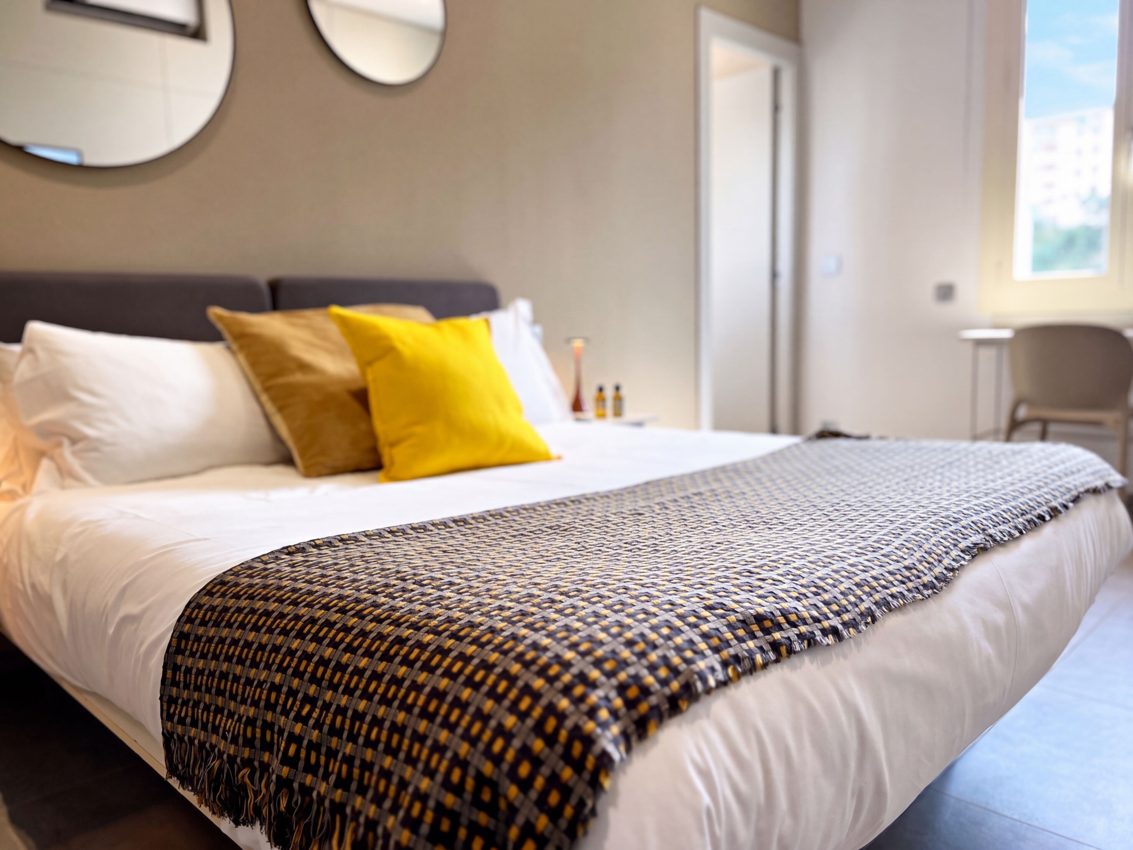A neatly made bed with white linens and a patterned throw blanket, a bedside table with a lamp, and a chair in a modern hotel room.