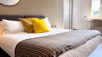 A neatly made bed with white linens and a patterned throw blanket, a bedside table with a lamp, and a chair in a modern hotel room.