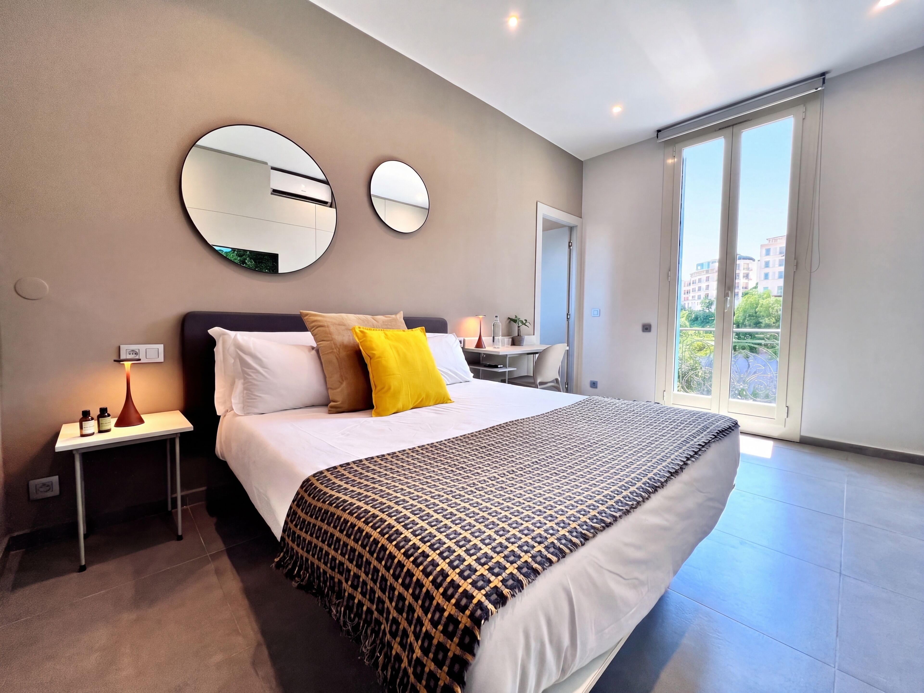 A modern hotel room with a large bed, two bedside tables, a desk, and a balcony with a city view.