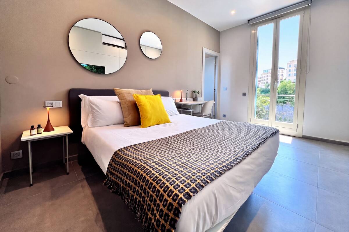 A modern hotel room with a large bed, two bedside tables, a desk, and a balcony with a city view.